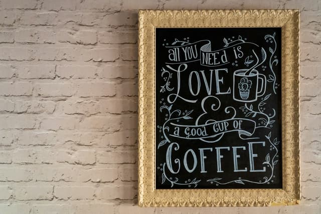 Framed hand-lettered chalk piece reading 'All you need is love & a good cup of coffee' on a white brick wall.